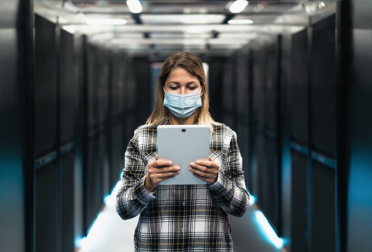 A woman wearing a face mask stands in a server room, holding and using a tablet.
