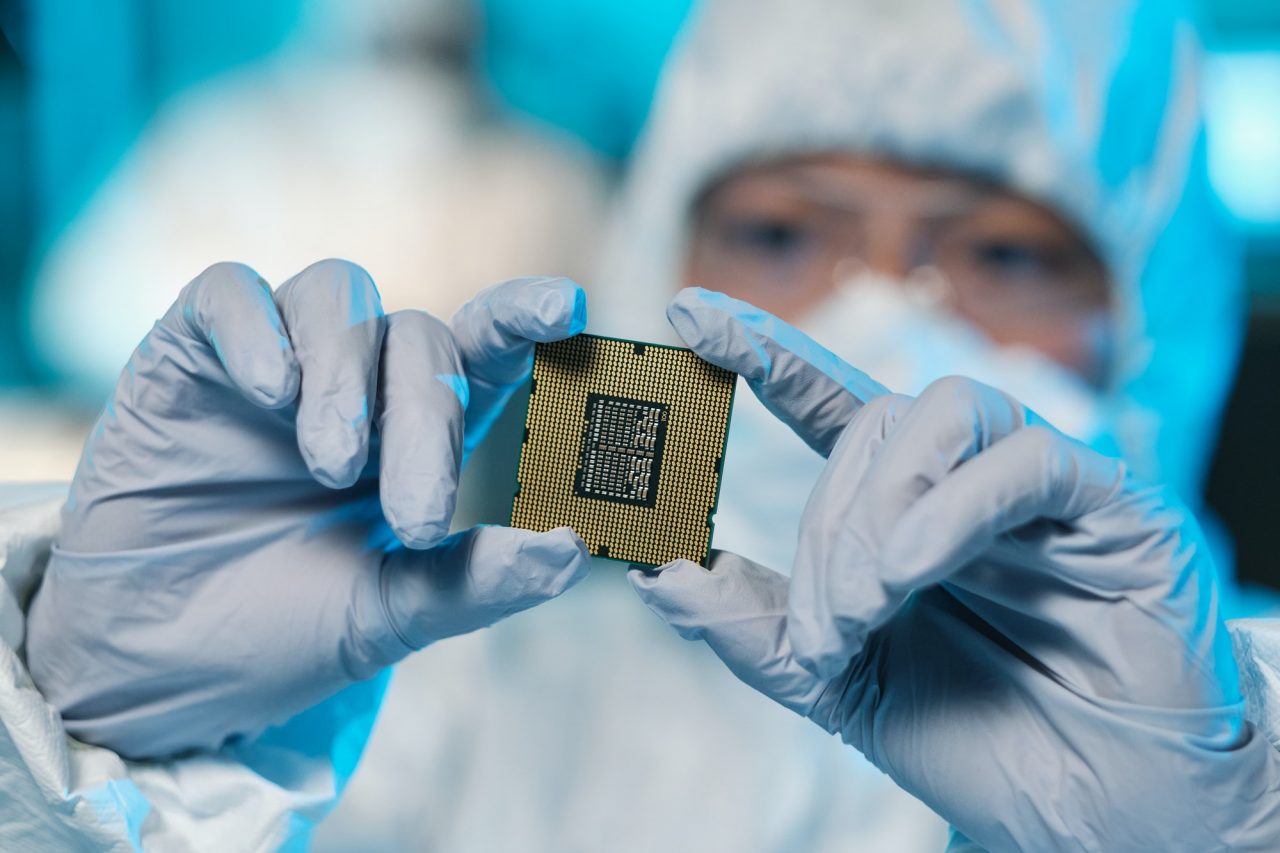 A person in protective gear holds a computer chip between gloved hands in a laboratory setting.