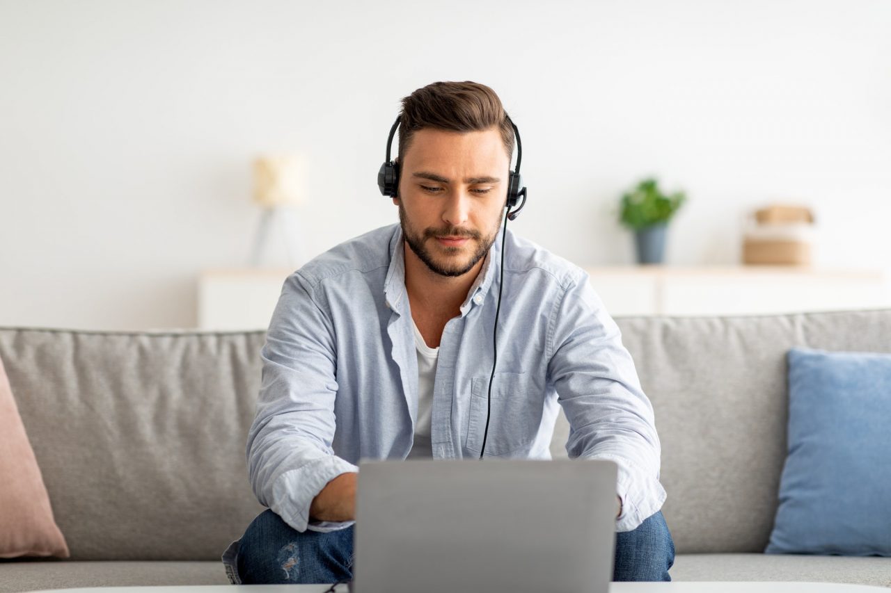 Man wearing a headset sits on a sofa and looks at a laptop screen, appearing focused. A blurred background shows a plant and home decor.