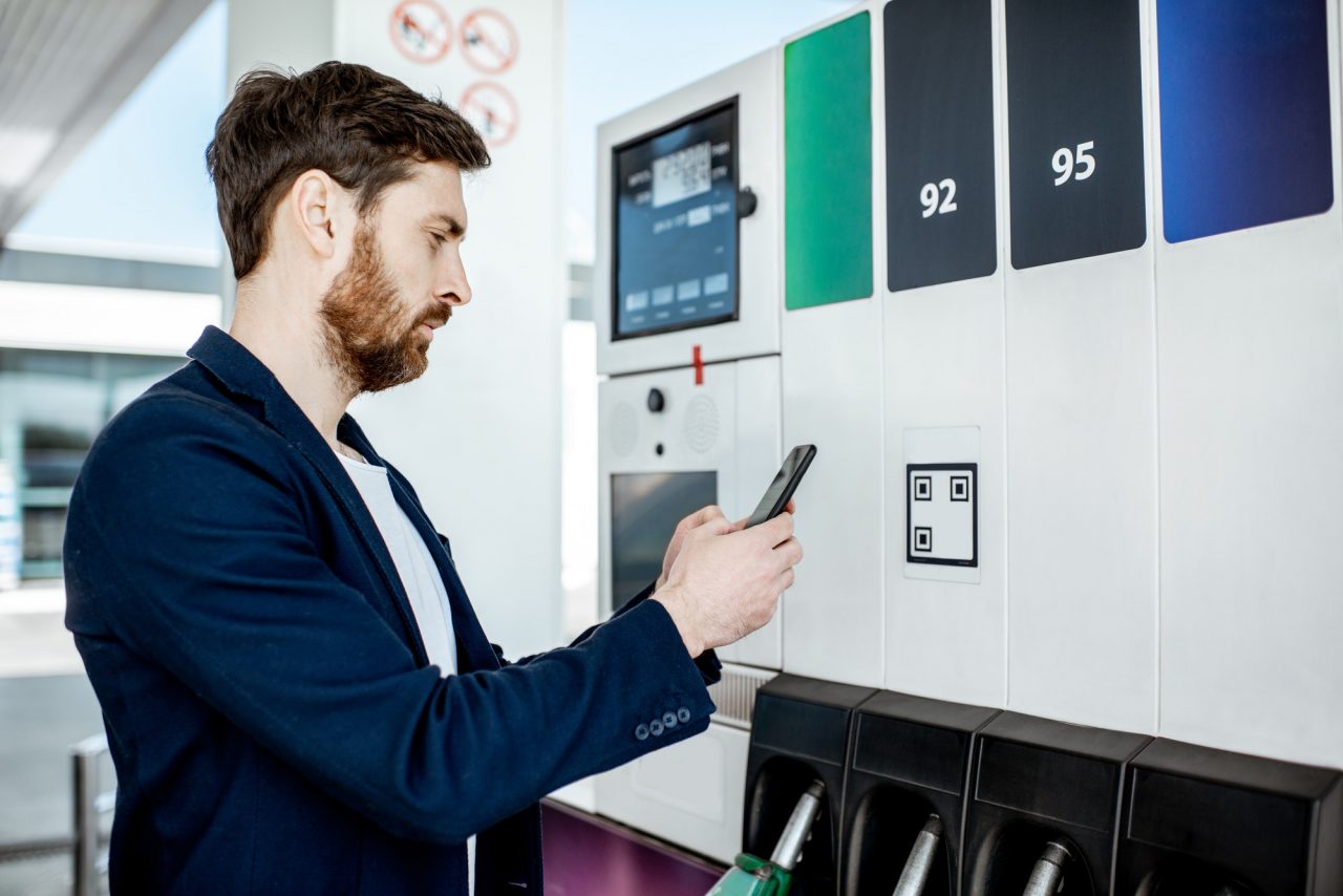 A man stands at a gas station pump, holding a phone and scanning a QR code near the fuel options.