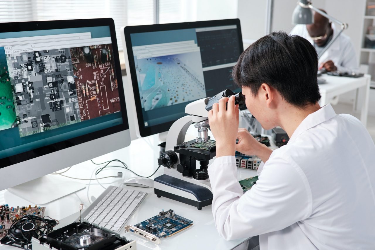A person in a lab coat examines a circuit board through a microscope at a desk with computer monitors displaying electronic component images.
