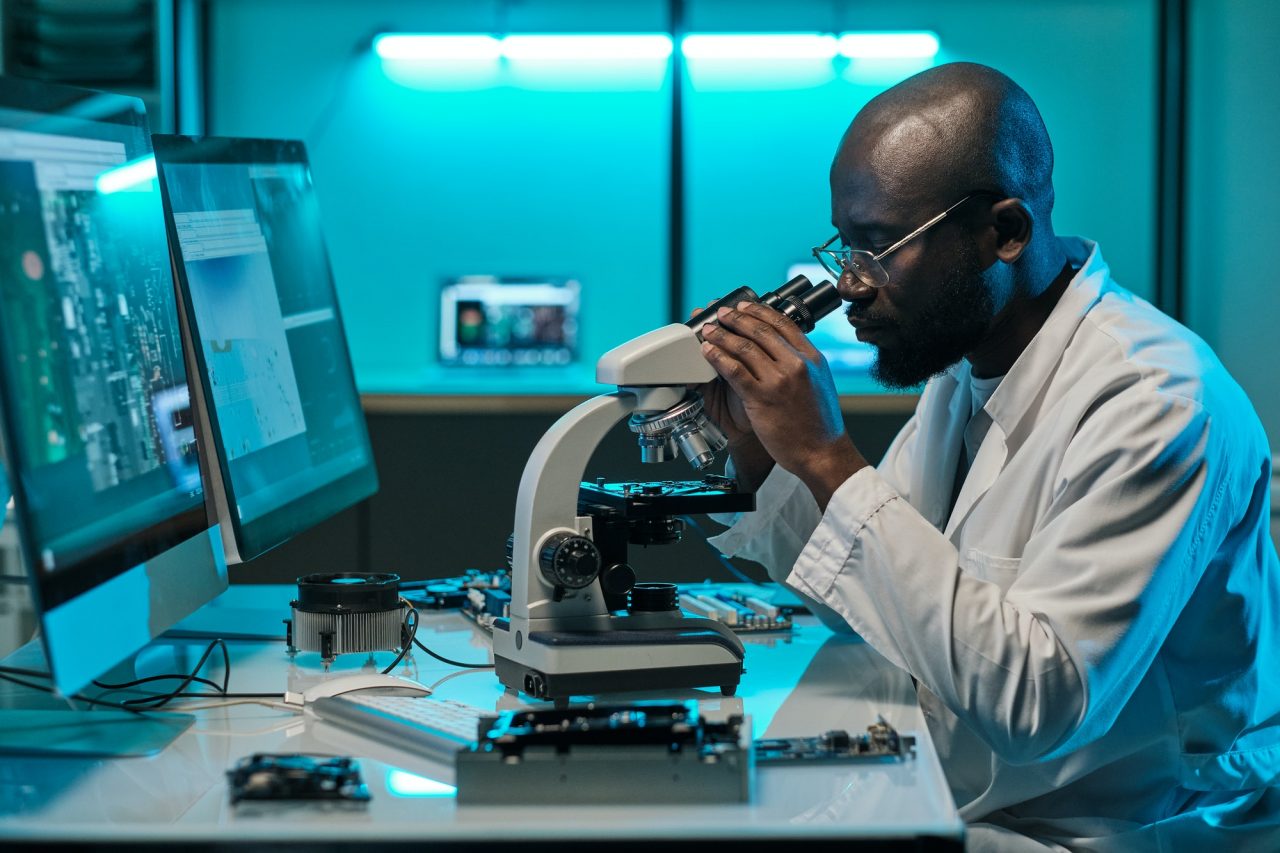 A scientist wearing a lab coat looks into a microscope at a workstation with computer monitors and electronic equipment in a laboratory setting.