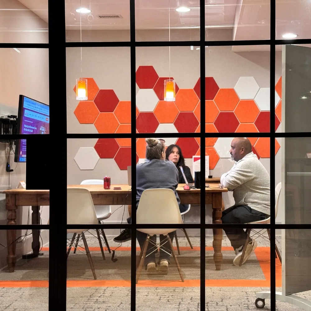 Three people sit at a conference table in a modern office meeting room with orange and red hexagonal wall decor, viewed through a glass window featuring a dynamic content widget display.