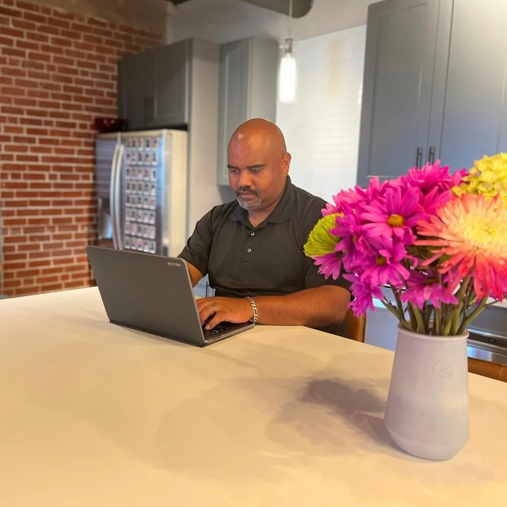 A man sits at a kitchen table working on a laptop, with a dynamic content widget open on his screen and a vase of colorful flowers in the foreground.