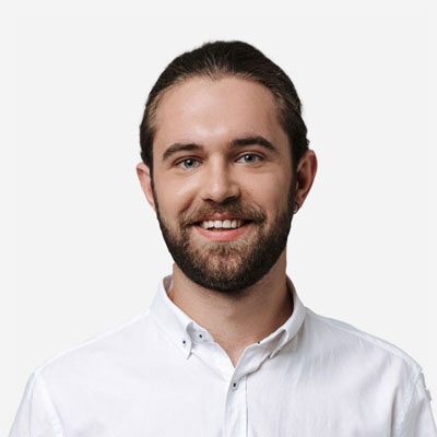 A man with brown hair and a beard, wearing a white button-up shirt, smiles at the camera against a plain white background.