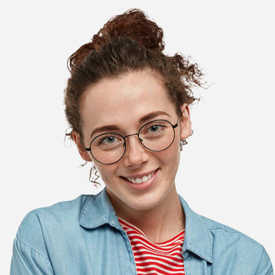 A young person with curly brown hair in a bun, wearing round glasses, a denim shirt over a red-and-white striped top, smiles at the camera against a plain background.