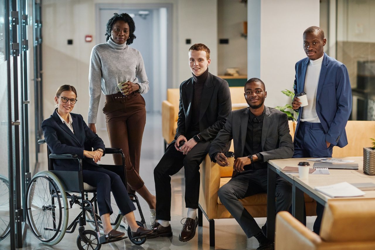 Five professionally dressed people, including one in a wheelchair, pose in a modern office setting with a laptop and notebooks on a nearby table.
